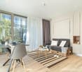 Spacious living room with large windows, gray sofa, wooden coffee table, striped rug, and a dining area with a table and chairs. The room has a modern and minimalist design with neutral colors and natural materials.