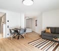Open-plan studio apartment with hardwood flooring, a dining table with chairs, a gray sofa, and a kitchenette with a dishwasher. The room is well-lit with a ceiling light fixture and has a minimalist, modern decor.