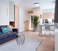 Open-plan studio apartment with living room, kitchen, and dining area. Hardwood floors, white walls, modern furniture including a navy blue sofa, coffee table, and desk with chair. Decorative plant and other accessories visible. Dishwasher can be seen in