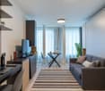 The image shows a modern and well-designed studio apartment. The main living area features a gray sofa, a striped rug, and a small dining table with a chair. The room has large windows that provide natural light and access to a balcony or terrace. The apa