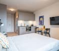 The image shows a studio apartment with an open-plan layout. The room features a bed, a small dining area with a table and chairs, and a kitchenette with cabinets, a stove, and a dishwasher. The walls are painted in neutral tones, and there is artwork and