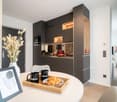 Modern kitchen with dark cabinets, built-in appliances, and a dining area with a round table and chairs. The kitchen features a dishwasher and is decorated with a vase of dried flowers and framed artwork on the wall.