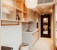 compact kitchen area with sink, oven, wooden cabinets, built-in shelves, woven basket, carpet, wooden side table, illuminated mirror