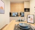 A modern dining area featuring a wooden table set for a meal. There are clear wine glasses, plates, cutlery, and a framed picture. Behind it, there is a kitchen with wooden cabinets, a black microwave, and a sink. A painting is on the wall.
