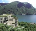 Modern multi-level house with balconies and terraces overlooking a scenic lake surrounded by lush, green mountains