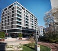 6-story building, gray and white facade, glass balconies, car parked nearby, garden area, statue, sign on the building, trees, bushes, street lights, clear blue sky, sunny