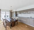 Dining room with wooden floor, large windows, dining table, chairs, chandelier, modern kitchen with gray cabinets, sink, dishwasher, and oven.