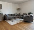 Spacious living room with hardwood floors, gray sofa and armchair, glass coffee table, and a floor lamp. The room has large windows, suggesting a balcony or terrace access.