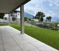 Spacious outdoor patio with tiled flooring, overlooking a lush garden with palm trees and a scenic mountain view in the background. The property appears to be a multi-story building with a balcony or terrace visible.