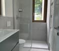 Modern bathroom with gray tile walls, glass shower enclosure, vanity with sink, and a window overlooking greenery outside.