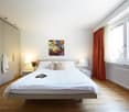 Spacious bedroom with a large bed, white bedding, and a modern artwork on the wall. The room has large windows with red curtains, providing natural light. There is a built-in wardrobe along one wall and a bedside table with lamps on either side of the bed