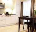 The image shows a modern kitchen and dining area. The kitchen features white cabinets, a dishwasher, and other appliances. There is a wooden dining table with chairs in the adjacent dining area. The room has large windows that provide natural light, and t