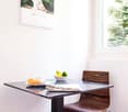 Dining table with chairs, bowl of fruit, a cup, croissant on a plate, magazine on the table, a framed art piece, large window with a view of trees