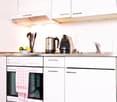 Modern white kitchen with built-in appliances, stainless steel countertop, built-in oven, toaster, coffee maker, kettle, bowl of fruits, cutlery set, faucet, sink, tiled flooring, cabinets