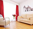 tan leather armchair, small table with flowers, cream couch with pillows, white wall, window with red curtains