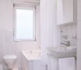 Bright and clean bathroom with white tiles, a toilet, a sink with a mirror cabinet, and a window providing natural light. A towel is hanging on the wall.