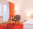 Spacious bedroom with large windows, orange curtains, wooden furniture including a chair and side tables, and a lamp. The room has a cozy and modern feel.