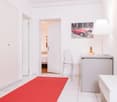 Bright and modern apartment with white walls, tile flooring, and a red carpet leading to the bedroom. The room features a desk with a lamp and a framed artwork of a red vintage car on the wall. The apartment appears to have a balcony or terrace, which is
