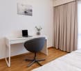 A white desk with a laptop, a plant, and a chair. An empty bed is present on the right side.