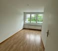 Empty room with white walls, large windows overlooking greenery, and hardwood flooring.