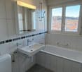 Spacious bathroom with large windows providing a scenic view, white tiles on the walls, a bathtub, and a sink with a mirror above it.