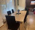 Dining room with large wooden table, black chairs, wooden flooring, white curtains on windows, table mat, and two bottles.