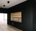 The image shows a modern, minimalist room with black walls and a wooden accent wall. The room features a built-in cabinet or entertainment unit with a wooden panel and a sink or appliance. The floor is made of light-colored hardwood. The room is illuminat