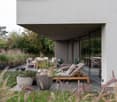 Gray house with glass walls, large lounge area with lounge chairs, plants, and dining table.