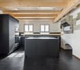 Black cabinets, concrete floor, island with gas stove, microwave and oven, wooden ceiling beams, white walls, stone fireplace