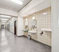 Hallway with tiled walls, ceiling lights, row of lockers, two sinks with mirrors and shelves, trash cans, and a window.