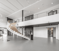 Large open-concept space with high ceilings, modern glass and steel staircase, white walls, and concrete flooring. The space appears to be a commercial or office building with multiple levels and glass doors leading to other areas.