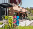 Brick building with a restaurant called 'Gus's Kitchen', outdoor seating area, colorful planters, and a modern architectural design with a canopy structure.