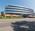 A multi-story office or commercial building with a curved, glass facade and red accents. The building has a modern, architectural design and is surrounded by trees and a paved area in front.