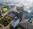 Cityscape with tall buildings, roads, greenery, open space, and vehicles. Central tall building with many windows, surrounded by smaller structures. Trees and a large green area visible in the center of the aerial view. Other visible buildings include apa