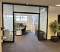 Office space with glass walls, desk, chairs, air conditioning, plants, and a trash bin.