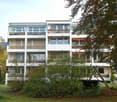 3-story apartment building with white exterior, large windows, and balconies. The building is surrounded by trees and greenery.