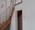 The image shows the interior of a building, with a wooden staircase and a metal railing system. The walls are painted white, and there is a wooden beam visible on the ceiling. The floor appears to be a dark-colored tile or concrete. There is a doorway vis