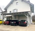 White two-story house with brown roof, three cars parked under carport, brick pavement