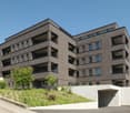 Multi-story apartment building with brick facade, balconies on each floor, and landscaped greenery surrounding the building.