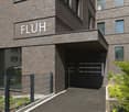 A multi-story brick building with the word 'FLUH' displayed prominently on the facade. The building has a garage entrance and is surrounded by greenery.