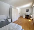 Single bed, nightstand, white lamp, white wardrobe, desk with black chair, desk lamp, picture frame, trash can, mirror, white walls, wooden floor
