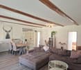 Living room with wooden beams, wooden floor, dining table, chairs, couch, coffee table, fireplace, shelves, picture, lamp, plants