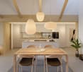 The image shows an open concept living space with a dining area and kitchen. The space features a wooden dining table with chairs, pendant lighting fixtures, and a modern kitchen with a central island and appliances. The walls are light-colored, and the c