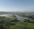 Aerial view of a scenic rural landscape with rolling hills, green fields, a winding river, and distant mountains in the background. The scene appears to be a picturesque countryside setting with a mix of agricultural and natural elements.