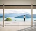 Large open-plan living space with floor-to-ceiling windows offering a stunning view of a lake surrounded by mountains. The room has a minimalist design with a white tiled floor, wooden ceiling panels, and white columns. A person is standing on the balcony