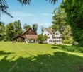Two-story white house with a wooden barn-like structure, surrounded by lush green trees and a well-manicured lawn. The property appears to have a balcony or terrace.