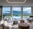 Spacious living room with large windows offering a panoramic view of the surrounding countryside and mountains. The room features a comfortable sectional sofa, a round wooden coffee table, and a plush armchair. The room also has access to an outdoor terra