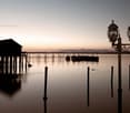 The image shows a serene lakeside or coastal scene at sunset. There is a wooden dock or pier extending into the calm, reflective water, with several wooden posts or pilings visible. In the background, there is a small building or structure silhouetted aga