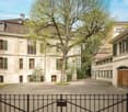 A historic multi-story building with a courtyard, surrounded by trees and other buildings. The building has a traditional European architectural style with arched windows, shutters, and a sloped roof. There is a wrought-iron gate in the foreground leading