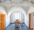 This image shows a vaulted ceiling room with ornate, decorative plasterwork. The room has a wooden table and chairs in the center, and the floor is covered with blue slate tiles. There are wooden doors on either side of the room, and a large arched window